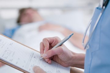 Photo of a nurse holding a patient's chart on a clipboard with a patient in bed in the background