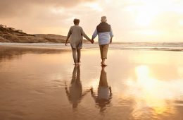 Two elderly people holding hands and walking on the beach as the sun sets