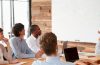 Photo of a woman standing at a white board as she leads a meeting and people gathered around a table look on