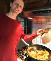 Person in red shirt standing in front of a cooktop holding a lid over a wok full of food