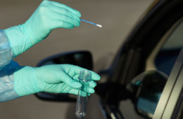 Photo of a gloved hand reaching out with a test tube and swab next to a car