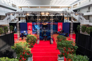 Photo of the courtyard of the Samson Pavilion with red carpet covering the floor, trees in planters surrounding the area and the debate stage with lights overheads