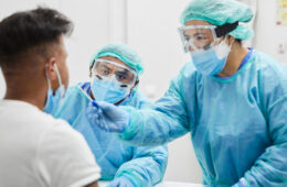 Photo of two health care professionals in protective gear giving a COVID swab test to a person